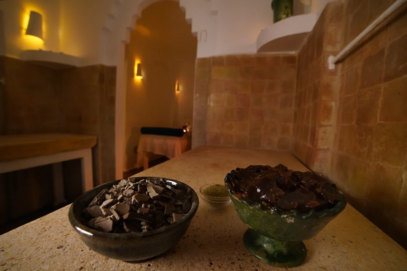 Cuencos de rhassoul y jabón negro sobre la encimera, con el pasillo del hammam al fondo